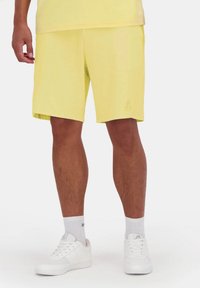 Pantalones cortos amarillos de tela ligera, con un corte relajado, bolsillos laterales y un logo sutil en el dobladillo. Combinados con zapatillas blancas.