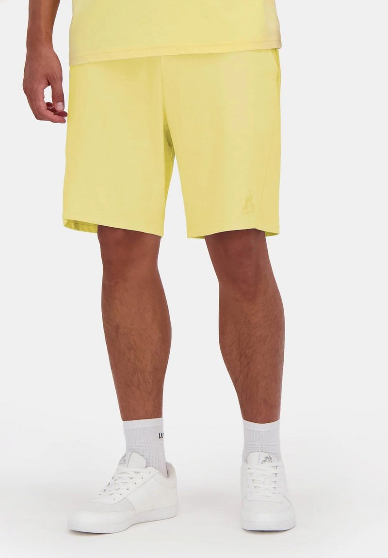 Pantalones cortos amarillos de tela ligera, con un corte relajado, bolsillos laterales y un logo sutil en el dobladillo. Combinados con zapatillas blancas.