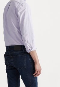 Muž v světle fialové dlouhé košili s ohrnutými manžetami a tmavě modrých džínsech, stojící na bílém pozadí, otočený zády.