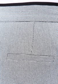 Kangasta, jossa on musta-valkoinen geometrinen kuvio, on vaakasuora tasku ja kontrastivärinen musta reunus reunoilla.