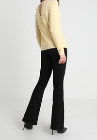 Pull jaune à dos ouvert avec manches longues, associé à un jean noir évasé. Le tissu semble doux et le design comporte un détail découpé dans le dos.
