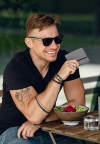 Černé tričko, krátké vlasy a sluneční brýle. Šedá peněženka v ruce. Miska s zeleným salátem s květinou na dřevěném stole. Sklenice s čistou vodou.