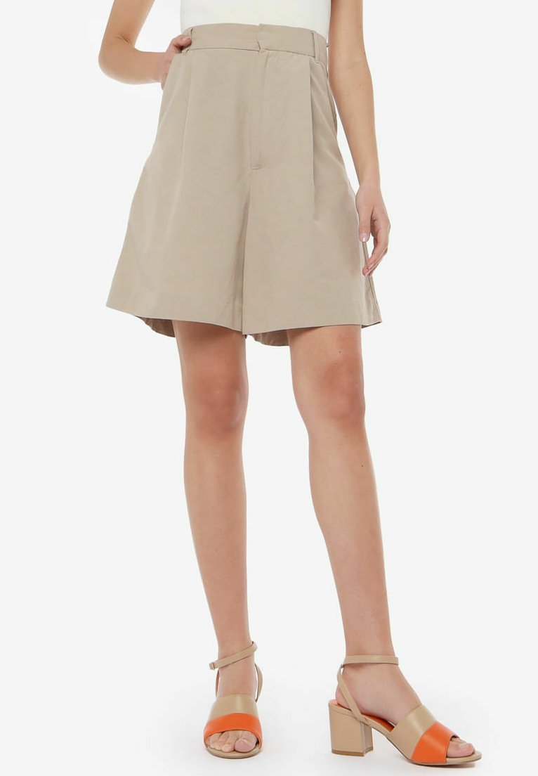 Pantalones cortos de talle alto en un tono beige claro con pliegues, hechos de una tela suave. Combinados con sandalias de tacón bloque en tonos marrón y naranja.