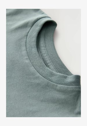 Trumpa rankovėmis marškiniai prislopintų žalių atspalvių, pasižymintys banguota apykakle ir lygiu tekstūru. Artimas vaizdas, akcentuojantis apykaklės detalę.