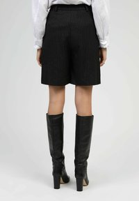 Shorts à rayures noires avec une coupe ajustée, dotés d'une ceinture. Associés à des bottes en cuir noires montantes et à un blouse blanche à manches longues.