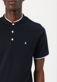 Navy Poloshirt mit Mandarin-Kragen, weißen Akzenten am Kragen und an den Ärmeln sowie vier weißen Knöpfen auf der Vorderseite mit einem Logo auf der Brust.