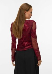 Kvinne med langt brunt hår iført en gjennomsiktig burgunder overdel med blomsterprint og svarte bukser, vendt bort fra kameraet mot en hvit bakgrunn.