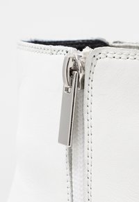 Botín de tobillo de imitación de cuero blanco con cremallera lateral de hardware metálico, costuras cosidas y un suave forro interior negro.