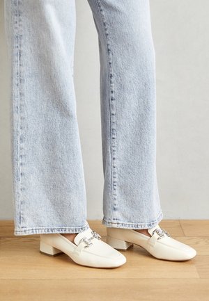 Mocassini in pelle bianca con tacco quadrato e punta squadrata, con dettagli in metallo argentato, abbinati a jeans a gamba larga blu chiaro.