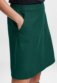Jupe verte avec une taille élastique et des poches latérales. Le tissu est lisse avec un léger éclat, présentant une coupe droite et un ourlet.