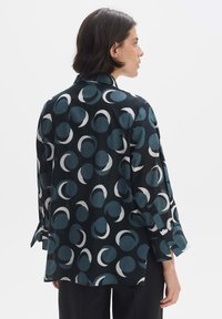 Camicia a maniche lunghe in tessuto scuro con grandi motivi circolari in teal e bianco. Presenta un colletto classico e polsini con bottoni. Vestibilità sagomata.