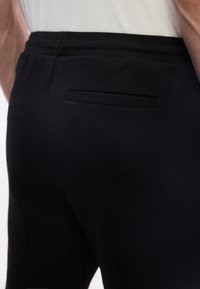 Πίσω όψη ενός ατόμου που φοράει μαύρο παντελόνι με μία οριζόντια πίσω τσέπη και λευκό τοπ.