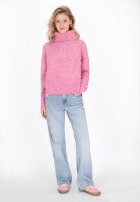 Fuchsia gestrickter Pullover mit hohem Kragen und geripptem Saum, kombiniert mit hellblauen weiten Jeans und rosa Schnürschuhen.