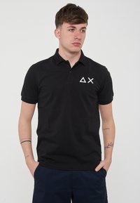 Polo nero in cotone. Presenta un colletto con bottoni, maniche corte e un logo geometrico bianco sul petto sinistro. Texture liscia.