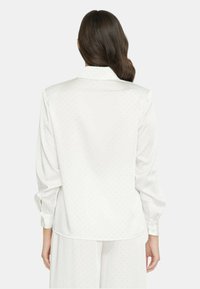 Camicia di seta bianca con maniche lunghe, colletto e piccole fantasie a pois. La vista posteriore mostra una texture liscia e una leggera lucentezza.