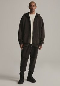 Ciemnobrązowy bawełniany hoodie i joggery o luźnym kroju, z zamkiem z przodu, dużymi kieszeniami i teksturowanymi akcentami, w połączeniu z czarnymi butami.