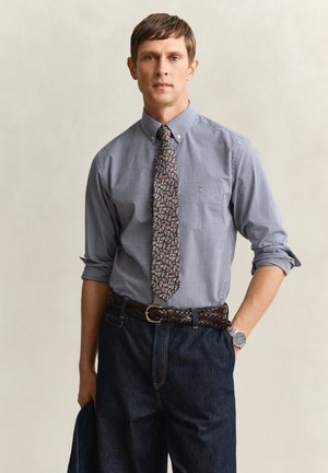 Man die een blauw geruit overhemd draagt met opgerolde mouwen, een stropdas met patroon, een gevlochten bruine riem, donkere spijkerbroek, en een marineblauw jasje vasthoudt.