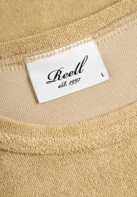 Beige Sweatshirtkragen aus einem strukturierten, weichen Stoff. Mit einem gewebten Etikett "Reell" und "est. 1997", Größe "L" auf einem weißen Hintergrund.