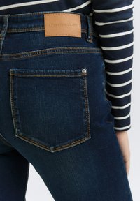 Jean en denim bleu foncé avec une étiquette en cuir portant une marque, des accents de couture et une poche arrière avec un détail à bouton-pression en argent.