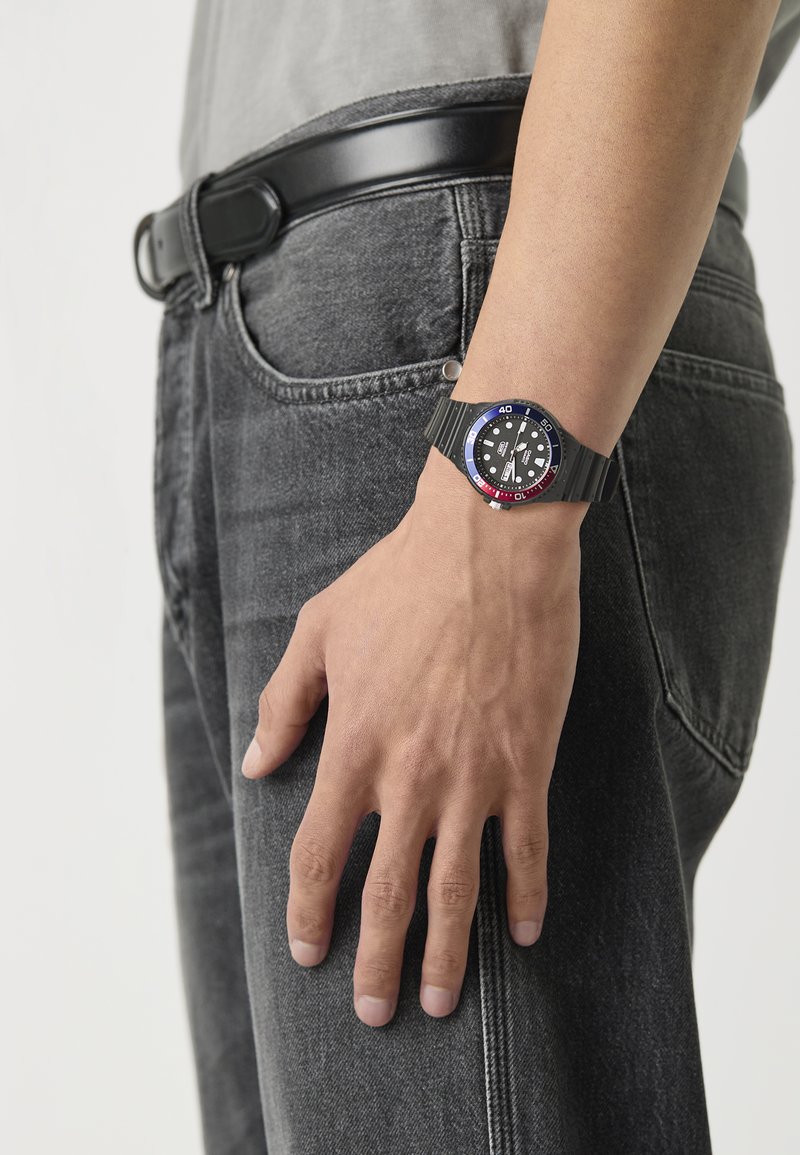 Montre de plongée noire et rouge avec un bracelet en caoutchouc, présentant un cadran rond, des index horaires blancs, et une lunette unidirectionnelle, posée sur un poignet à côté d'un jean gris.