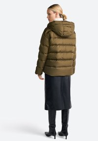 Doudoune vert olive avec capuche, présentant des sections matelassées et une texture lisse. Associée à une jupe midi en cuir noir et des bottes à talons.