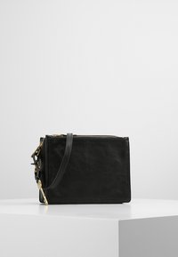 Sort læder crossbody taske med lynlås og gyldne detaljer, placeret på en hvid pedestal mod en minimalistisk baggrund.