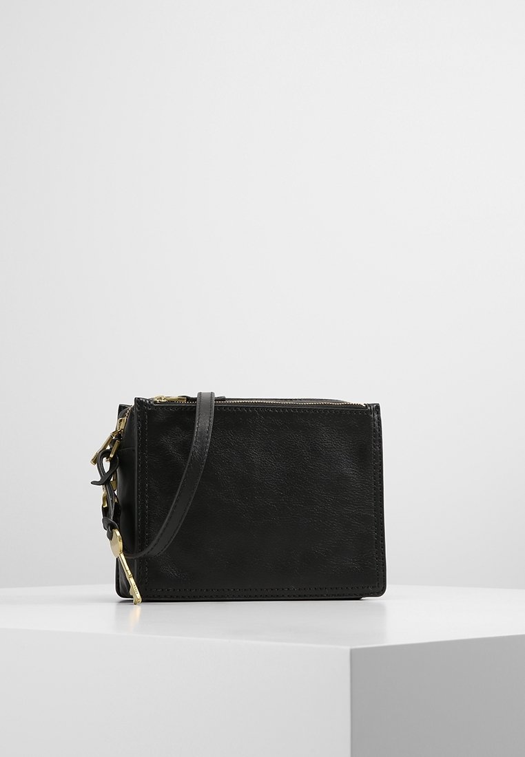 Sort læder crossbody taske med lynlås og gyldne detaljer, placeret på en hvid pedestal mod en minimalistisk baggrund.