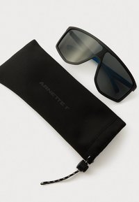 Occhiali da sole neri Arnett con bracci interni blu accanto a un astuccio in tessuto morbido nero con chiusura a cordoncino su una superficie bianca.