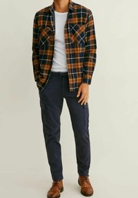 Man står klädd i en brun och marinrutig skjorta över en vit t-shirt, marinblå cargobyxor och bruna läderstövlar, med avslappnade händer vid sidorna.