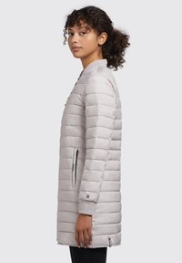 Cămașă puffer gri deschis, matlasată, cu guler înalt, buzunare cu fermoar și manșete ribbate. Are un design elegant și prelung pentru căldură.