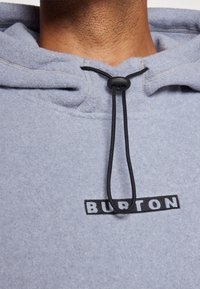 Pilkas flisinis džemperis su juodais raišteliais ir reguliavimo mazgu. Priekyje matomas juodas "BURTON" logotipas. Minkšta, tekstūruota medžiaga arti.