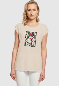 Beige bomulds T-shirt med korte ærmer, prydet med et Frida Kahlo grafik i livlige farver og fed sort skrift. Enkel, afslappet pasform.