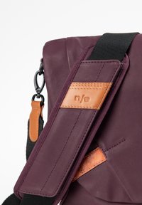 Borsa di stoffa viola con tracolla nera regolabile, rifiniture in pelle marrone sulla tracolla e sulla borsa, e chiusura a clip in metallo su sfondo bianco.