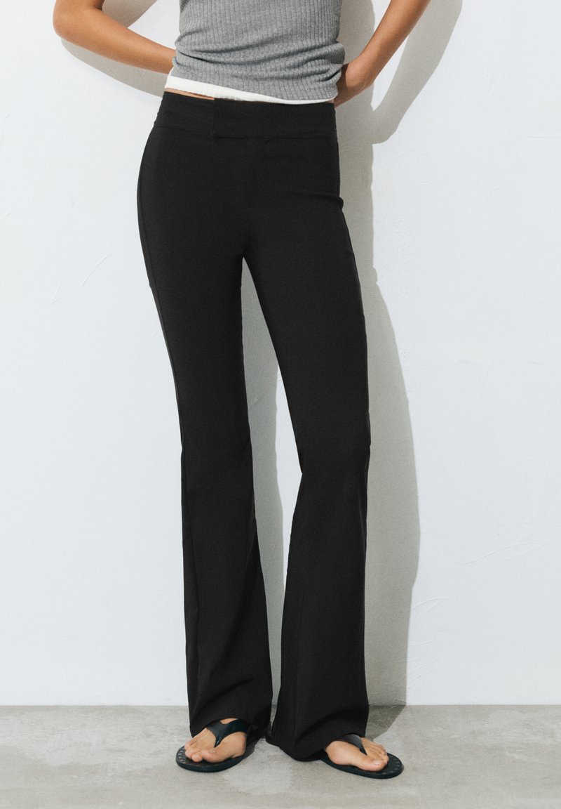 Femme portant un pantalon noir évasé et des tongs noires, debout contre un mur blanc, mains posées sur les hanches.