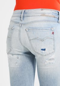 Light blue distressed denim jeans featuring frayed edges and multiple rips. Back pockets have a distinct design and red stitching accent.
