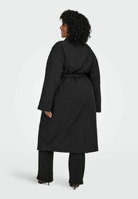 Melns garš mētis ar jostas vidukli un plašām piedurknēm. Audums izskatās gluds ar smalku spīdumu. Stāvot pret gaismas fona.