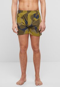 Zelena in rjava patterned kopalke z elastičnim pasom in kratko dolžino, ki vključujejo geometrijske oblike v mornarsko modri in khaki barvi.