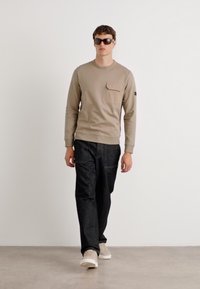 Sweatshirt bege claro com um bolso, combinado com calças de ganga escuras e ténis beges. Modelo a usar óculos de sol escuros, de pé contra um fundo claro.