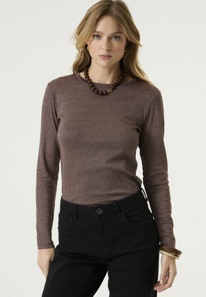 Frau mit blondem Haar, die ein braunes Langarmshirt, schwarze Hose, Perlenkette und Armbänder trägt, steht vor einem schlichten Hintergrund.