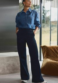 Cămașă din denim cu butoni, decorată cu accente negre, purtată împreună cu pantaloni din căptușeală de culoare bleumarin, cu talie înaltă, și adidași albi, pe un fundal modern.