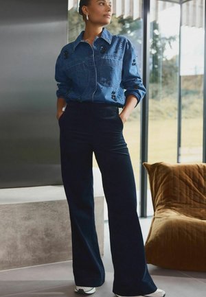 Cămașă din denim cu butoni, decorată cu accente negre, purtată împreună cu pantaloni din căptușeală de culoare bleumarin, cu talie înaltă, și adidași albi, pe un fundal modern.