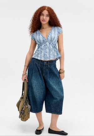 Giovane donna con capelli rossi e ricci indossa un top blu fantasia e pantaloni capri in denim larghi, tenendo una grande borsa marrone in una mano.