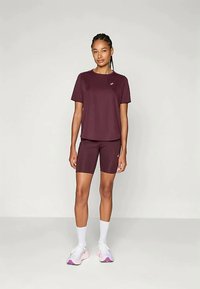 Nainen seisoo, päällään samansävyinen viininpunainen urheilupaita ja -shortsit, valkoiset sukat sekä vaaleanpunaiset urheilukengät, neutraali tausta.