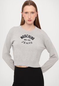 Izgriezta pelēka džemperis, kas izgatavots no mīksta materiāla, ar melnu adītu logotipu "MOSCHINO JEANS" un ribotu apdari apkaklē un apakšmalā.