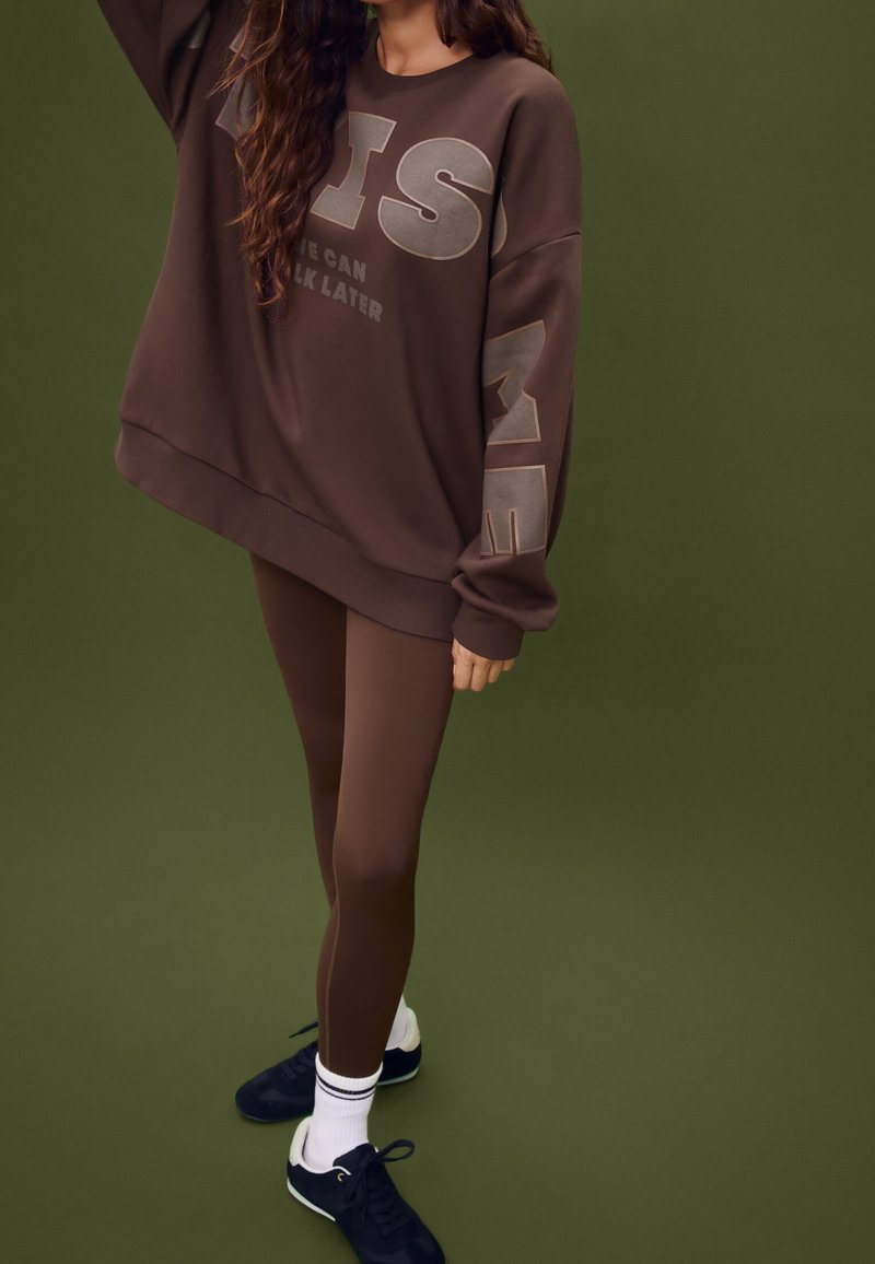 Personne portant un sweat-shirt marron oversize avec texte et leggings assortis, chaussettes blanches et baskets noires sur fond vert olive.
