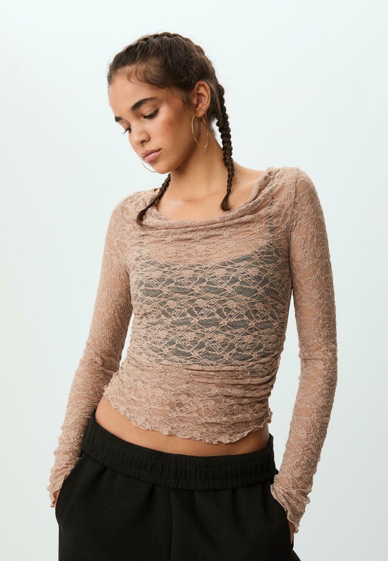 Lace langærmet top i lys beige. Har en draperet halsudskæring, struktureret blomstermønster og tætsiddende silhuet, parret med sorte bukser med talje.