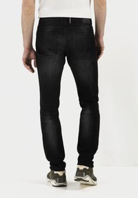 Schwarze Denim-Jeans mit schmaler Passform, ausgestattet mit Gesäßtaschen und einer mittelhohen Taille. Getragen mit grauen und weißen Sneakers, vor einem schlichten Hintergrund.