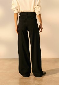 Pantaloni negri, cu picior larg și talie înaltă, cu două buzunare pe spate și cusături curate, pe un fundal neutru. Textura moale este evidentă.