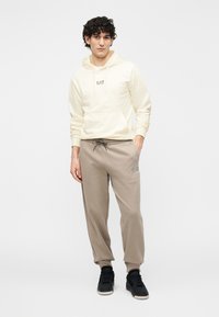 Jasnożółta bluza z kapturem ze sznurkiem, z małym logo. W zestawie beżowe spodnie dresowe i czarne buty sportowe. Gładka tekstura materiału.