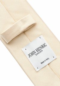 Beige tygslips med diagonala ränder, utrustad med en vit etikett som inkluderar "JOHN HENRIC," "SVERIGE," och "Tillverkad i Italien." Strukturerad yta.